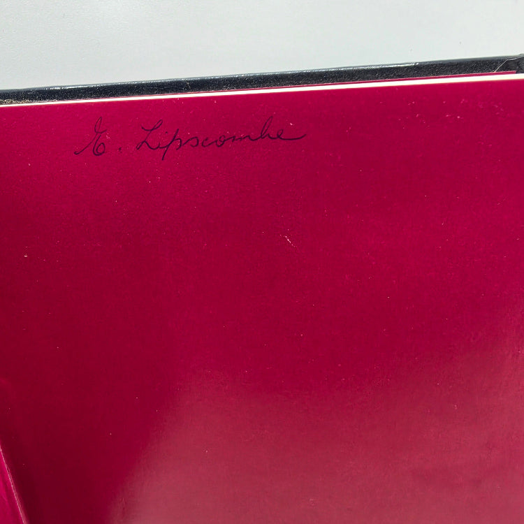A close-up of a glossy deep magenta-red surface with a small black handwritten signature near the top, against a light gray background.