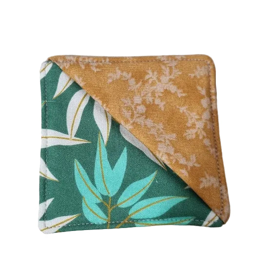 A square fabric bookmark with a diagonal split design, featuring a green side with white and turquoise leaf patterns and a mustard-gold side with white floral motifs.