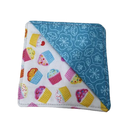 A square fabric bookmark with a diagonal split design. One side is white with colourful cupcake prints, and the other side is light blue with white snowflake and leaf patterns.