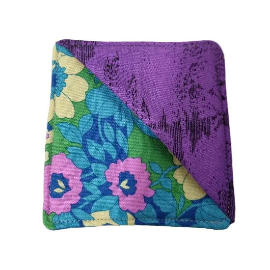 A fabric bookmark with a diagonal split design: one side is solid purple with a subtle dark pattern, and the other side features a retro floral print with pink, blue, green, and yellow flowers.