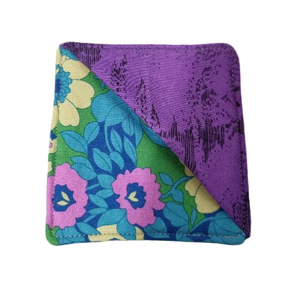 A fabric bookmark with a diagonal split design: one side is solid purple with a subtle dark pattern, and the other side features a retro floral print with pink, blue, green, and yellow flowers.