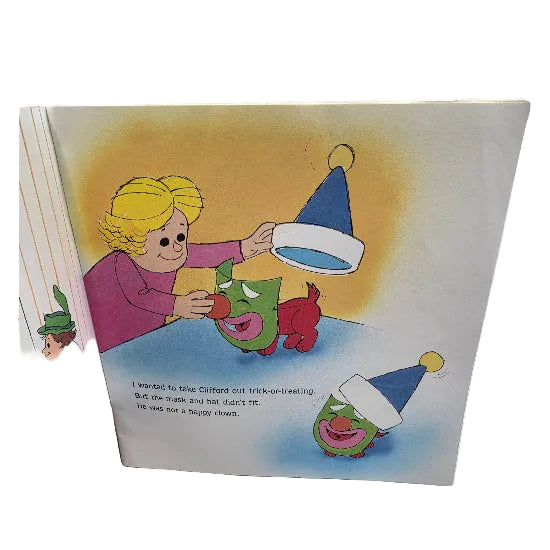 A page from Clifford's First Halloween showing a colourful illustrated page with a child helping Clifford, who is wearing a green mask, put on a blue party hat. The page includes printed text and is photographed against a plain white background.