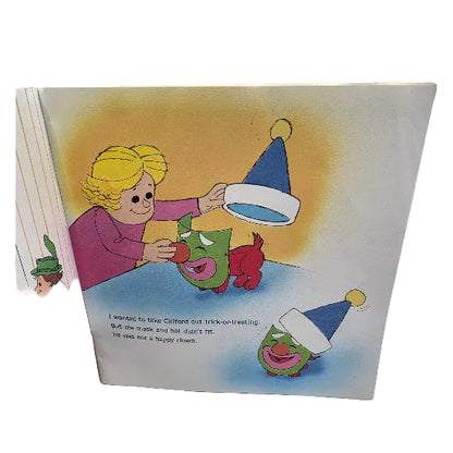 A page from Clifford's First Halloween showing a colourful illustrated page with a child helping Clifford, who is wearing a green mask, put on a blue party hat. The page includes printed text and is photographed against a plain white background.