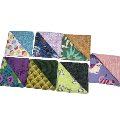 Seven square fabric bookmarks  arranged in two rows. Each piece has a split design with two contrasting patterned halves, featuring prints such as floral, butterfly, leaf, scallop, lamp, and cat motifs in multiple colours.
