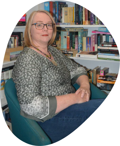 An image of Dorothy, a blonde woman wearing glasses and a floral shirt, sitting in a blue chair with bookshelves behind her. 