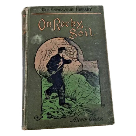 Vintage hardcover book titled 'On Rocky Soil' by Annie Gray, featuring an illustrated cover with a man breaking a large rock.