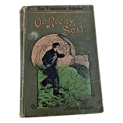 Vintage hardcover book titled 'On Rocky Soil' by Annie Gray, featuring an illustrated cover with a man breaking a large rock.